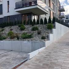 Nowoczesny taras z kostki brukowej prowadzący wzdłuż betonowego muru oporowego, z nasadzeniami roślinności na skalniaku i budynkiem mieszkalnym w tle.