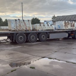 WORLDGRAN LOGISTICS SPÓŁKA Z OGRANICZONĄ ODPOWIEDZIALNOŚCIĄ - Ciężarówka z naczepą przewozi duże bloki kamienne zabezpieczone pasami. W tle budynki i firmowy bus transportowy. Mokry plac z odbiciami.