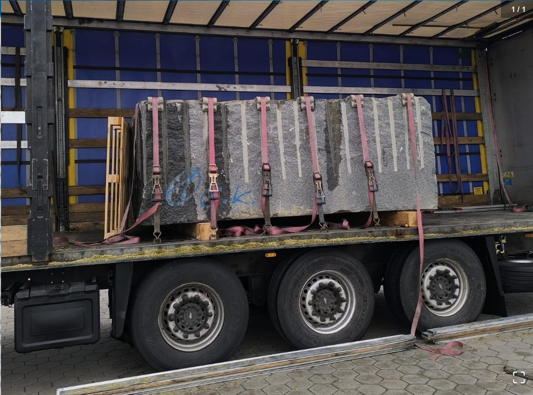 Ciężarówka z otwartą naczepą przewozi zabezpieczone pasami bloki kamienne na paletach, widoczne koła i fragment plandeki. Transport specjalistyczny.