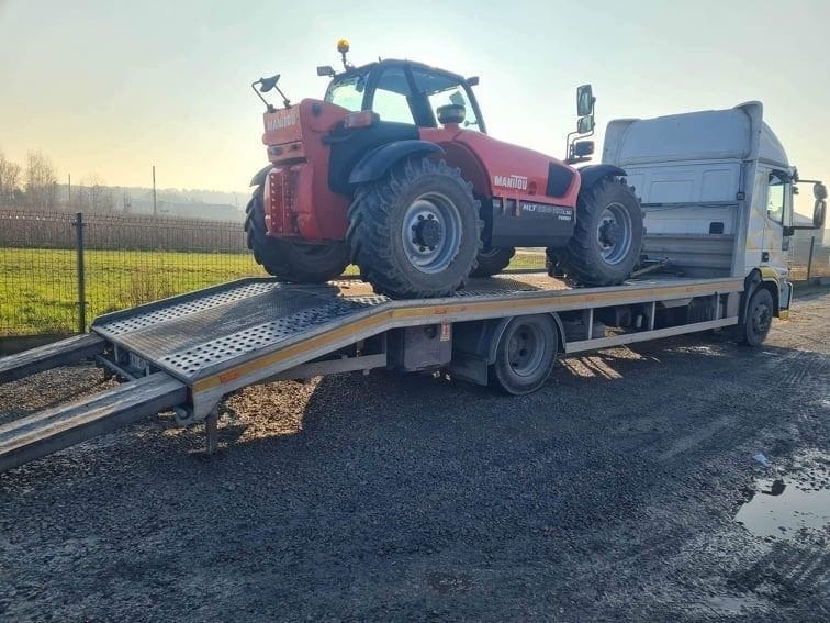 Czerwony wózek widłowy Manitou na platformie lawety transportowej, gotowy do przewozu. Widok z boku, słoneczny dzień.