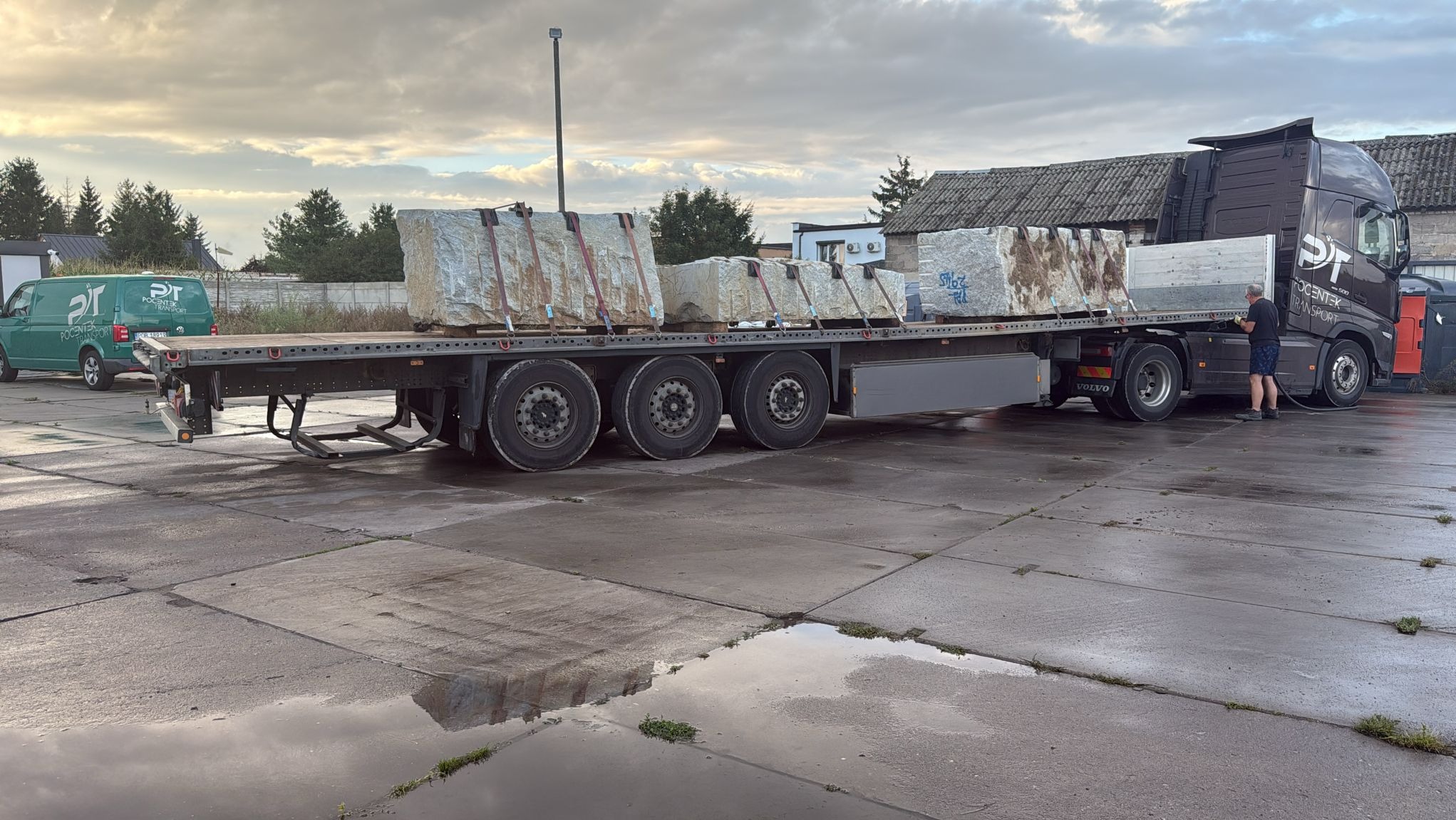 Ciężarówka z naczepą przewozi duże bloki kamienne zabezpieczone pasami. W tle budynki i firmowy bus transportowy. Mokry plac z odbiciami.