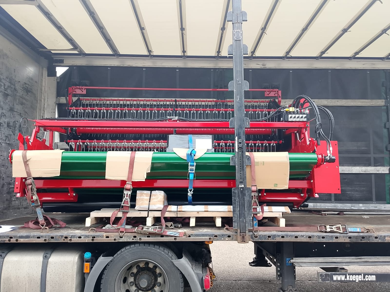 Czerwono-zielona maszyna rolnicza zabezpieczona pasami wewnątrz naczepy ciężarówki, widok z boku, częściowo zakryte koło i podwozie pojazdu.