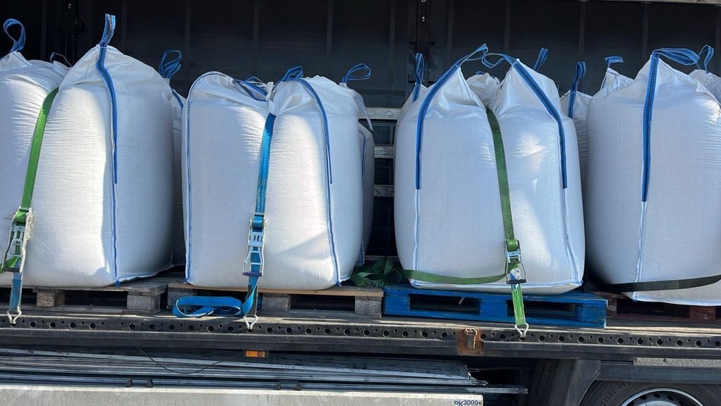 Załadunek worków typu Big Bag na paletach w przestrzeni ładunkowej pojazdu ciężarowego, zabezpieczonych pasami transportowymi. Widok z boku, jasne oświetlenie.