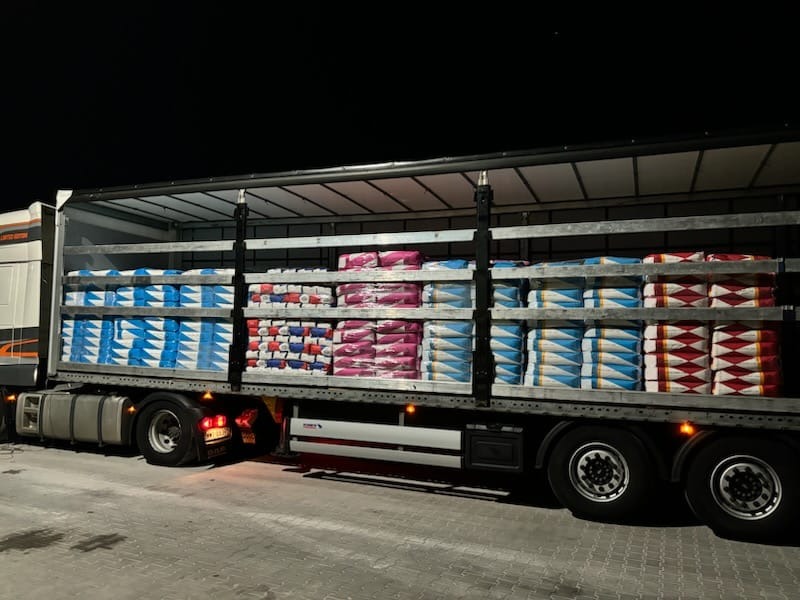 Ciężarówka z naczepą, załadowana paletami z workami w kolorowych opakowaniach, stojąca na parkingu nocą. Widok z boku, oświetlenie parkingu.