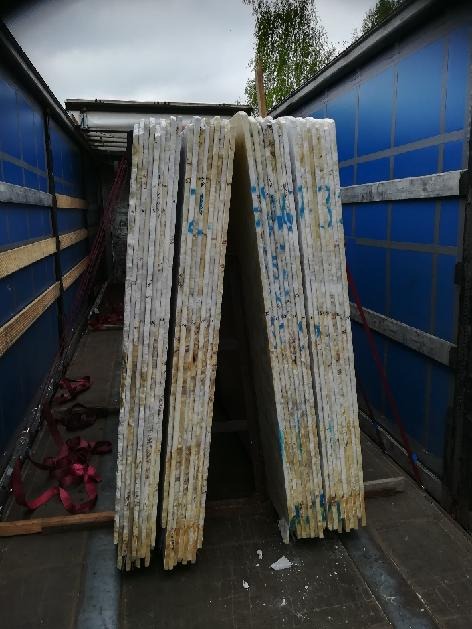 Paczki białych płyt z niebieskimi oznaczeniami, zabezpieczone wewnątrz niebieskiej naczepy ciężarówki, gotowe do transportu. Widoczne pasy zabezpieczające.