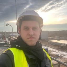 Oleh Vyshar - Elektryk w kasku i kamizelce odblaskowej na tle instalacji przemysłowej w trakcie budowy, widok z góry, zachodzące słońce.