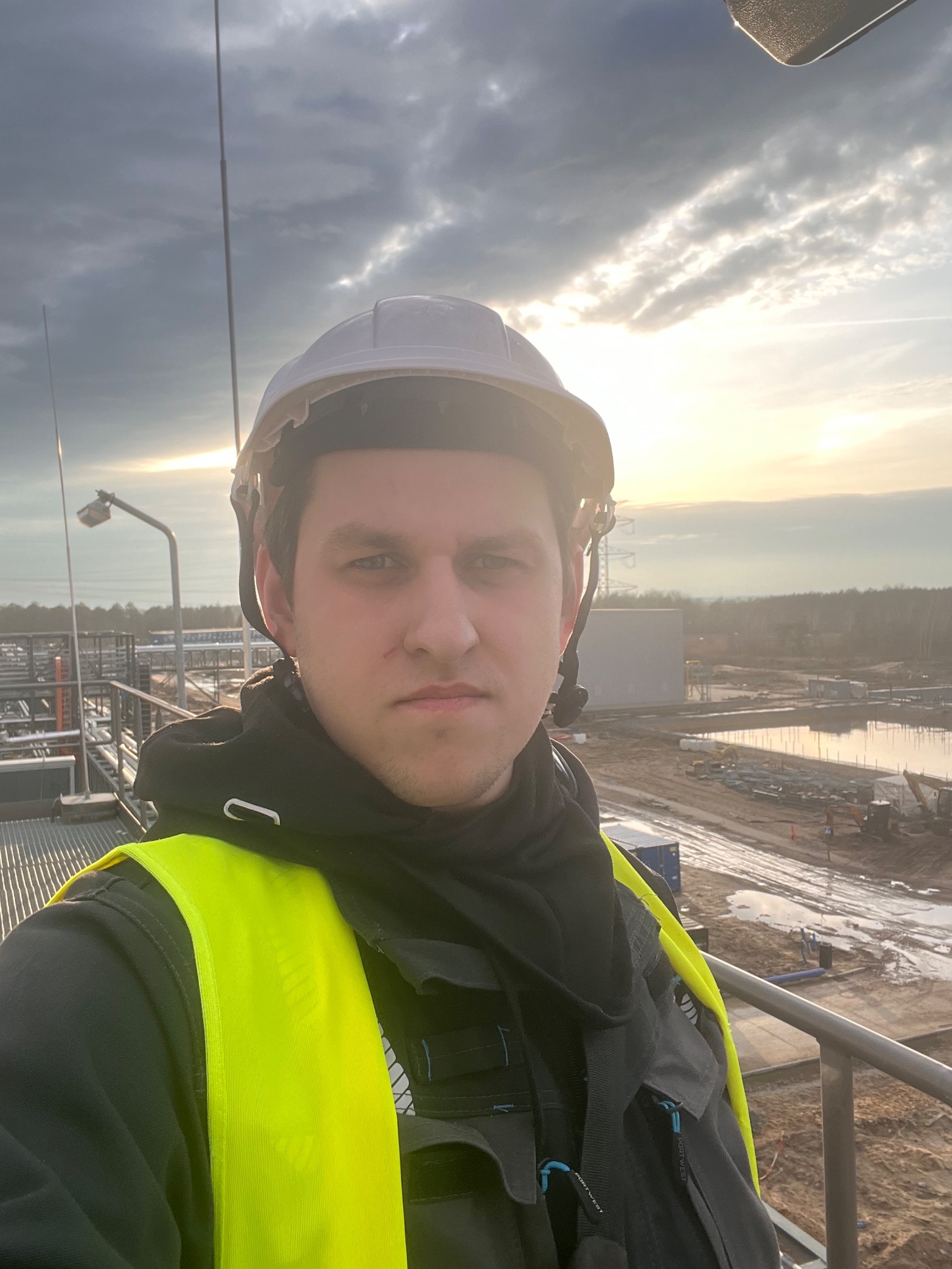 Elektryk w kasku i kamizelce odblaskowej na tle instalacji przemysłowej w trakcie budowy, widok z góry, zachodzące słońce.