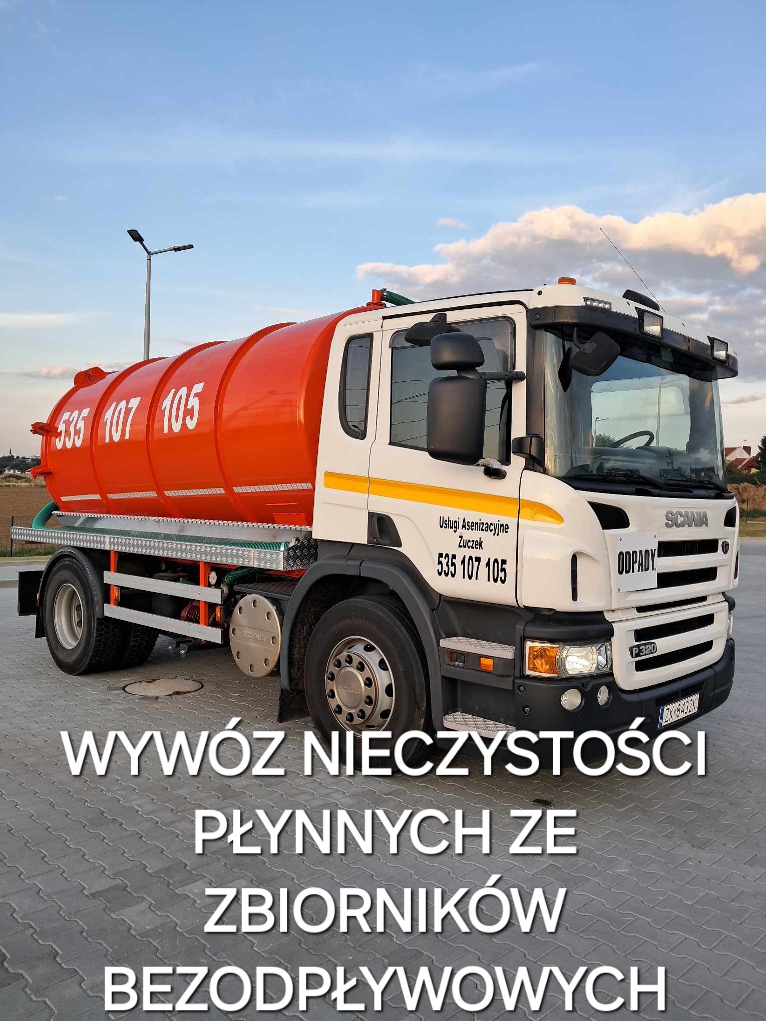 Ciężarówka asenizacyjna Scania z pomarańczową cysterną i numerem telefonu 535 107 105 na tle błękitnego nieba z chmurami, zaparkowana na betonowej nawierzchni.