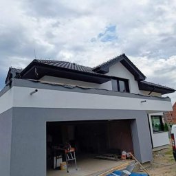 ProfiPBG - Dwukondygnacyjny dom z garażem w trakcie malowania elewacji na szaro i biało, widoczny otwarty garaż z narzędziami i drabina, na drugim planie inny budynek.