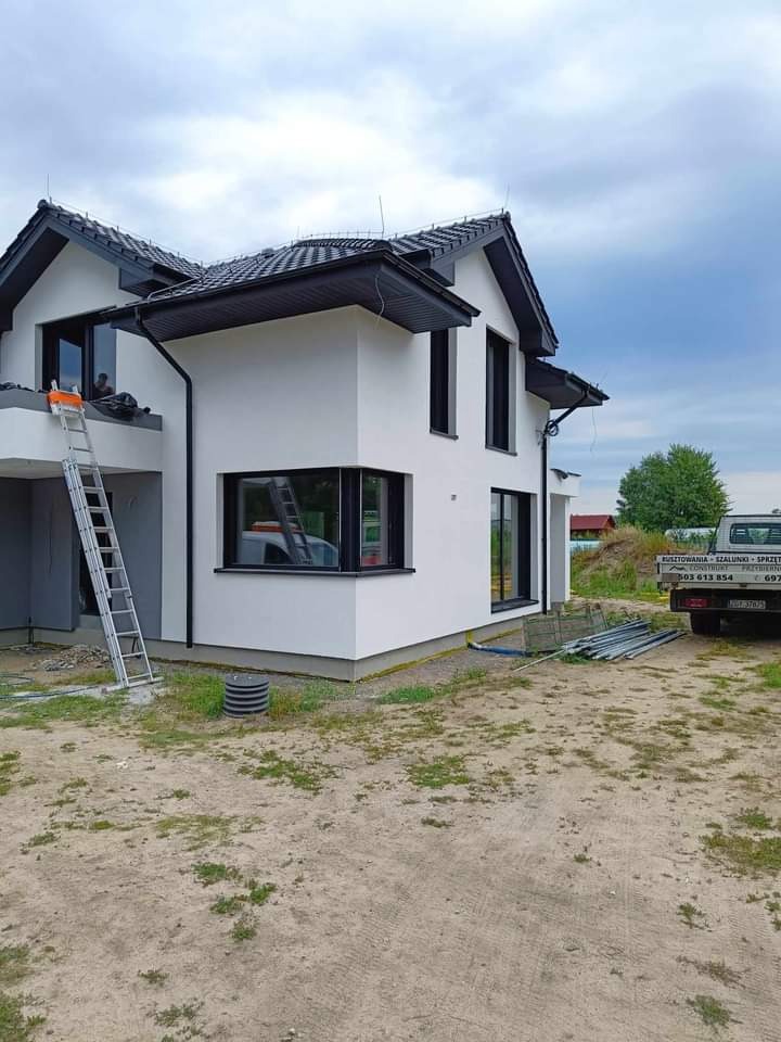 Nowoczesny dom jednorodzinny z białą elewacją, czarnymi ramami okiennymi i dachem, widoczna drabina oparta o ścianę, teren wokół domu częściowo zagospodarowany, w tle samochód dostawczy...