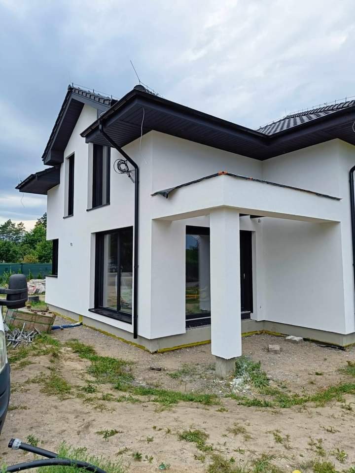 Nowoczesny, dwukondygnacyjny dom z białą elewacją i czarnymi ramami okiennymi, widok z zewnątrz, teren wokół domu częściowo zagospodarowany, widoczne ślady prac budowlanych.