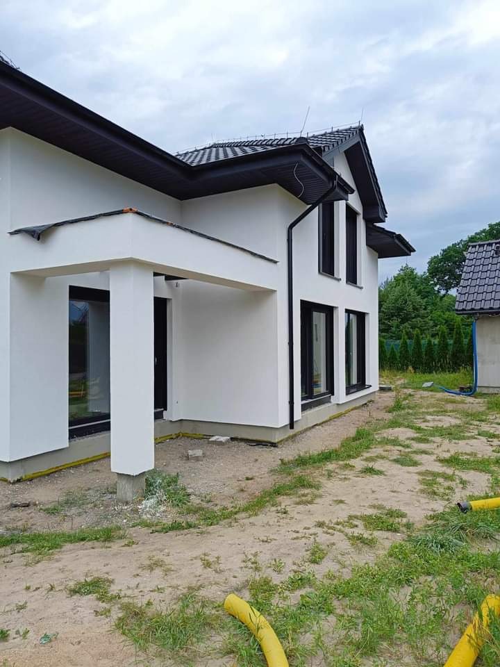 Nowoczesny dom z białą elewacją i czarnymi ramami okiennymi, widoczne żółte rury na trawniku, w tle drugi budynek z czarnym dachem