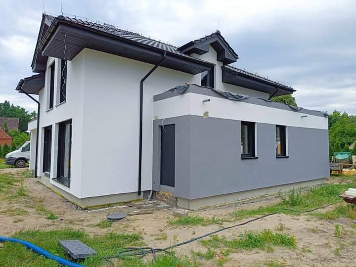 Dwukondygnacyjny dom z elewacją w odcieniach bieli i szarości, z czarnymi ramami okiennymi i dachem, widoczny niebieski wąż na trawniku przed budynkiem.