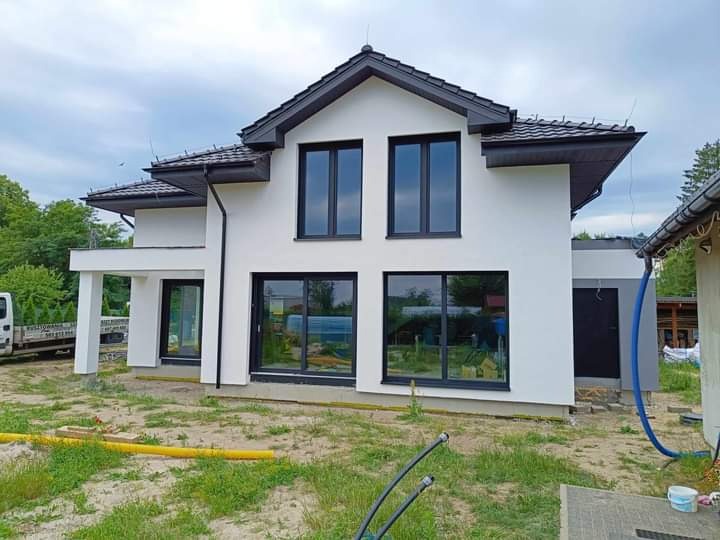 Nowoczesny, dwukondygnacyjny dom z białymi ścianami i ciemnymi oknami, otoczony zielenią, z widocznymi elementami instalacji wokół budynku.