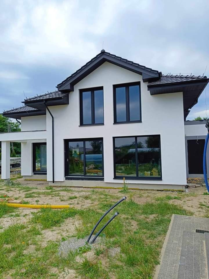 Nowoczesny, dwukondygnacyjny dom z białą elewacją, czarnymi oknami i dachem z ciemnej dachówki, widok z poziomu gruntu, z widocznymi wystającymi rurami w trawie.