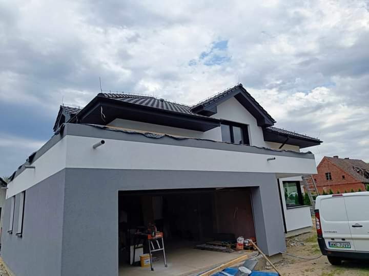 Dwukondygnacyjny dom z garażem w trakcie malowania elewacji na szaro i biało, widoczny otwarty garaż z narzędziami i drabina, na drugim planie inny budynek.