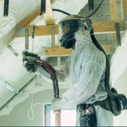 Pracownik w kombinezonie ochronnym aplikuje izolację natryskową na poddaszu, widoczne drewniane elementy konstrukcyjne i fragmenty świeżo nałożonej piany.