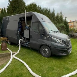 Ociepleniapoddaszy.pl - Szary samochód dostawczy z otwartymi drzwiami, wyposażony w sprzęt do natrysku pianki PUR, z widocznymi białymi wężami na trawniku prowadzącymi w kierunku budynku.