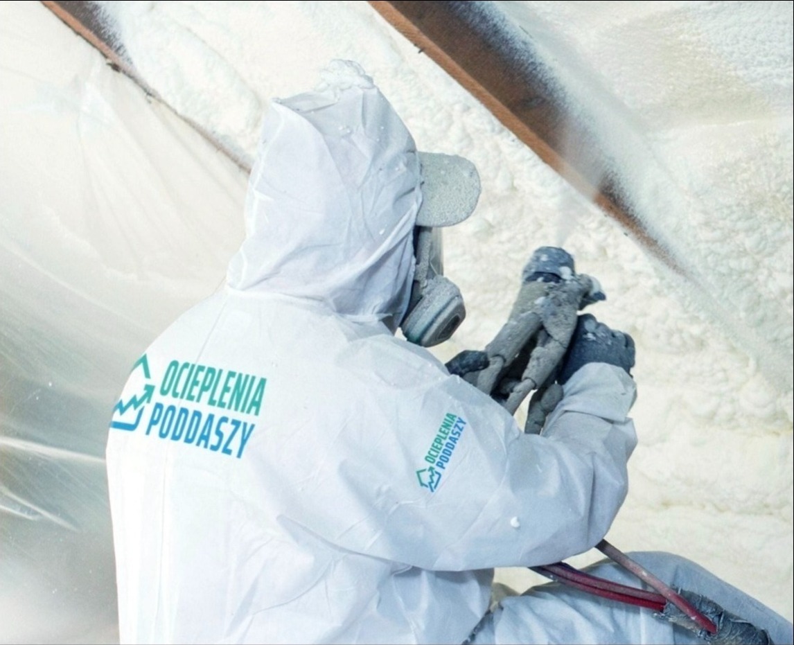 Pracownik w kombinezonie ochronnym z logo 'OCIEPLENIAPODDASZY.PL' natryskuje piankę PUR na poddasze, widoczne elementy konstrukcji drewnianej i świeżo nałożona izolacja.