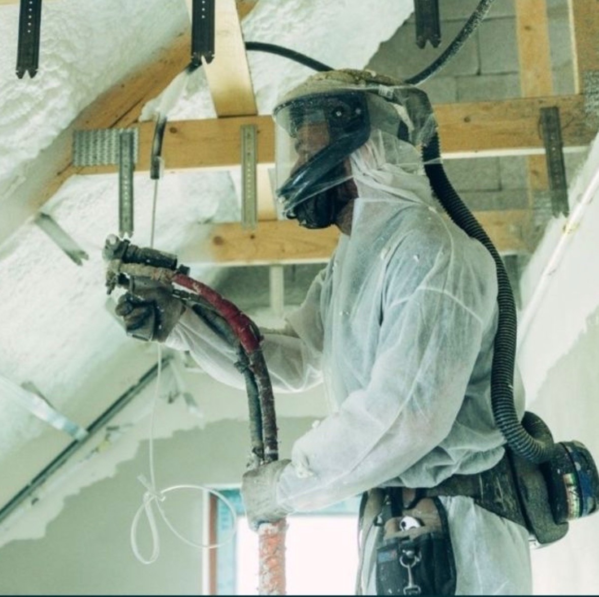 Pracownik w kombinezonie ochronnym aplikuje izolację natryskową na poddaszu, widoczne drewniane elementy konstrukcyjne i fragmenty świeżo nałożonej piany.