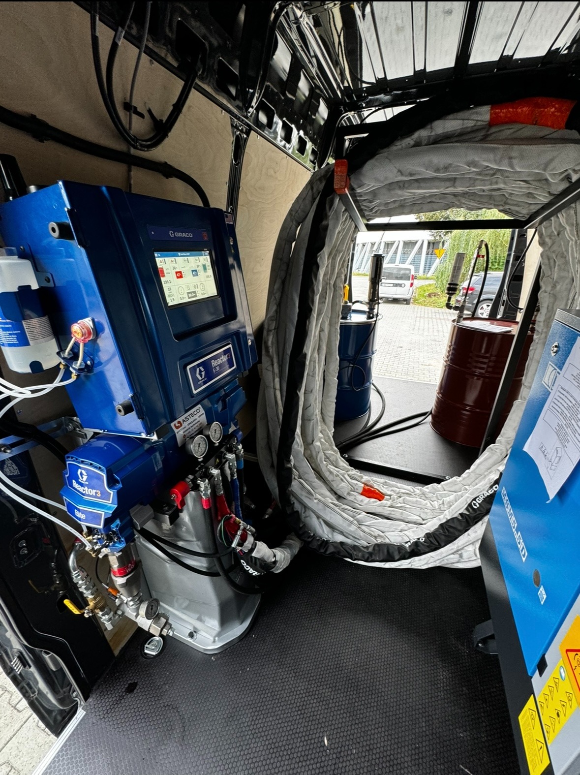 Wnętrze furgonetki z profesjonalnym agregatem natryskowym marki Graco, przeznaczonym do aplikacji pianki izolacyjnej, widoczne beczki z komponentami i zwinięte węże.
