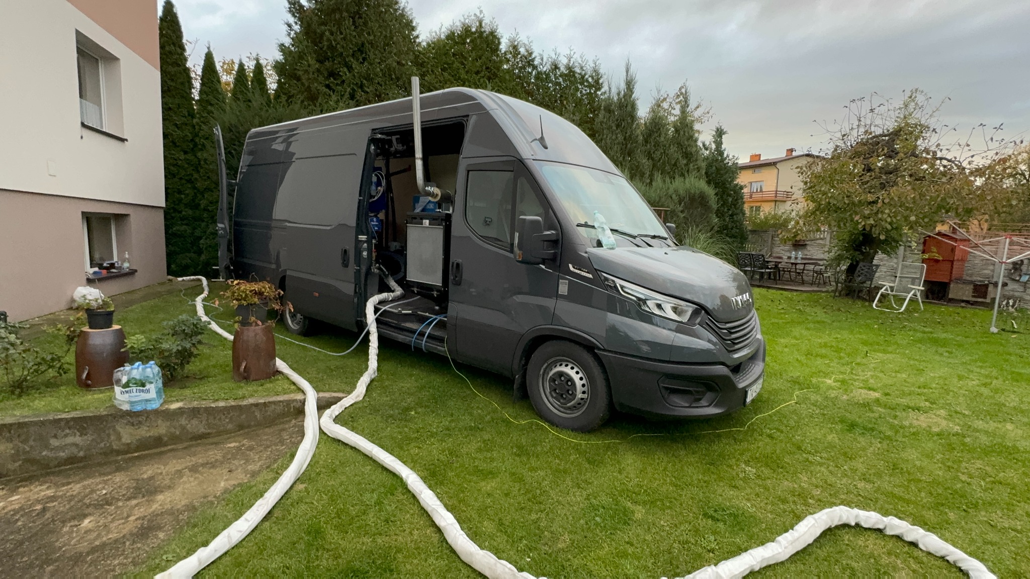 Szary samochód dostawczy z otwartymi drzwiami, wyposażony w sprzęt do natrysku pianki PUR, z widocznymi białymi wężami na trawniku prowadzącymi w kierunku budynku.