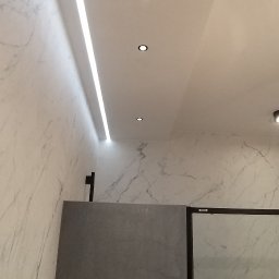 Top Bud Tomas Caputa - Nowoczesna łazienka z marmurowymi ścianami, oświetleniem LED i punktowymi lampami sufitowymi, widoczny fragment szklanej kabiny prysznicowej.