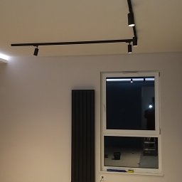 Top Bud Tomas Caputa - Minimalistyczny salon z czarnym grzejnikiem panelowym, oświetleniem szynowym na suficie i białymi ścianami, widok przez okno na zewnątrz.