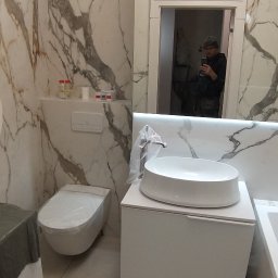 Top Bud Tomas Caputa - Nowoczesna łazienka z marmurowymi płytkami, owalną umywalką na szafce i toaletą w folii ochronnej, odbicie osoby robiącej zdjęcie w lustrze.