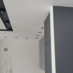 Top Bud Tomas Caputa - Szary pokój z oświetleniem sufitowym: czarna listwa z reflektorami, rząd okrągłych halogenów i wiszący kabel z żarówką.