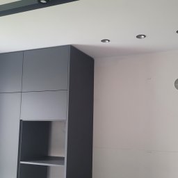 Top Bud Tomas Caputa - Szara zabudowa meblowa z otwartą półką, w tle biała ściana z widocznymi śladami po pracach wykończeniowych i punktowe oświetlenie sufitowe.