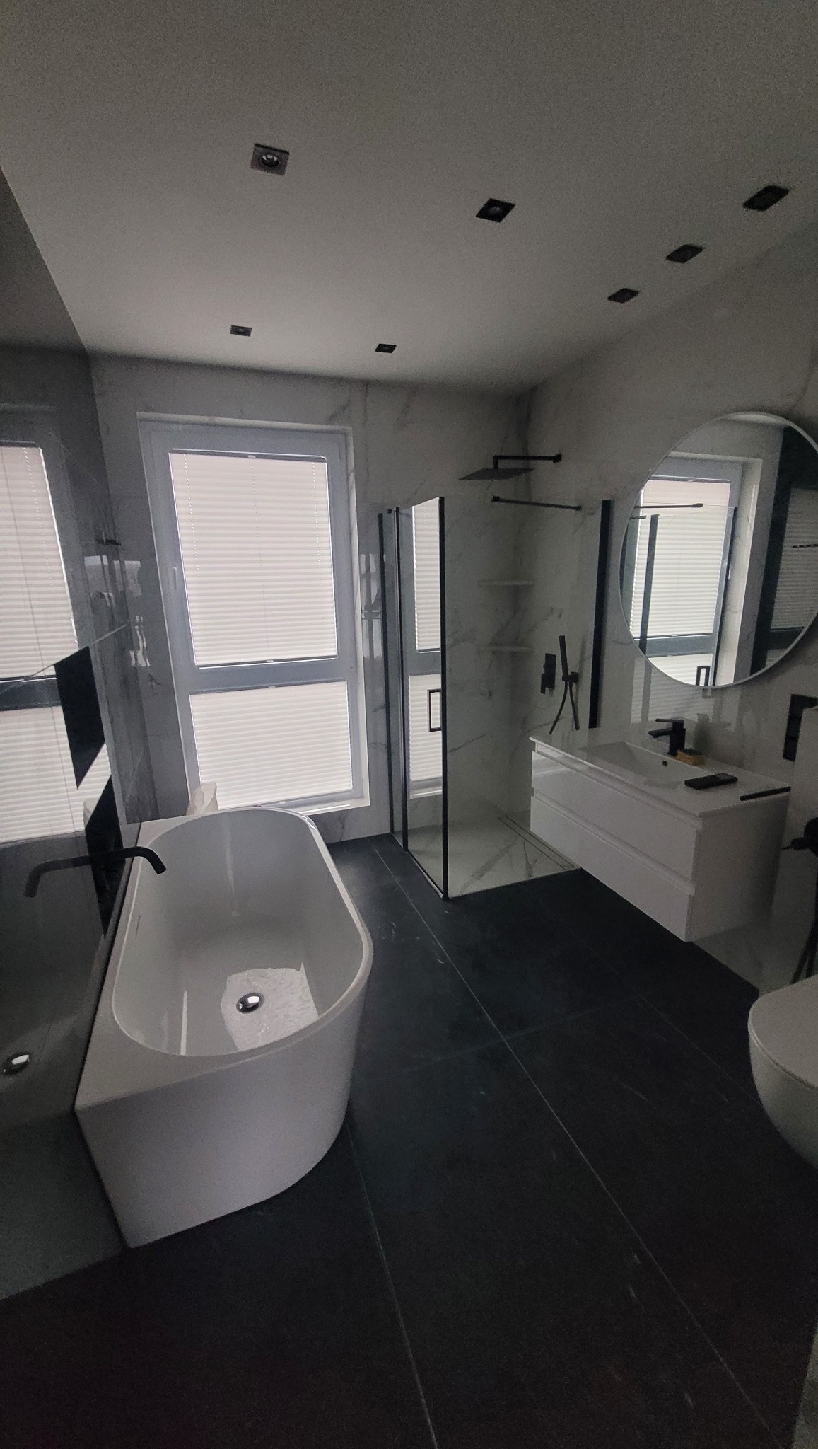 Nowoczesna łazienka z białą wanną wolnostojącą, kabiną prysznicową typu walk-in z czarnymi ramami, umywalką nablatową i czarną armaturą, wykończona marmurowymi płytkami i ciemną podłogą.