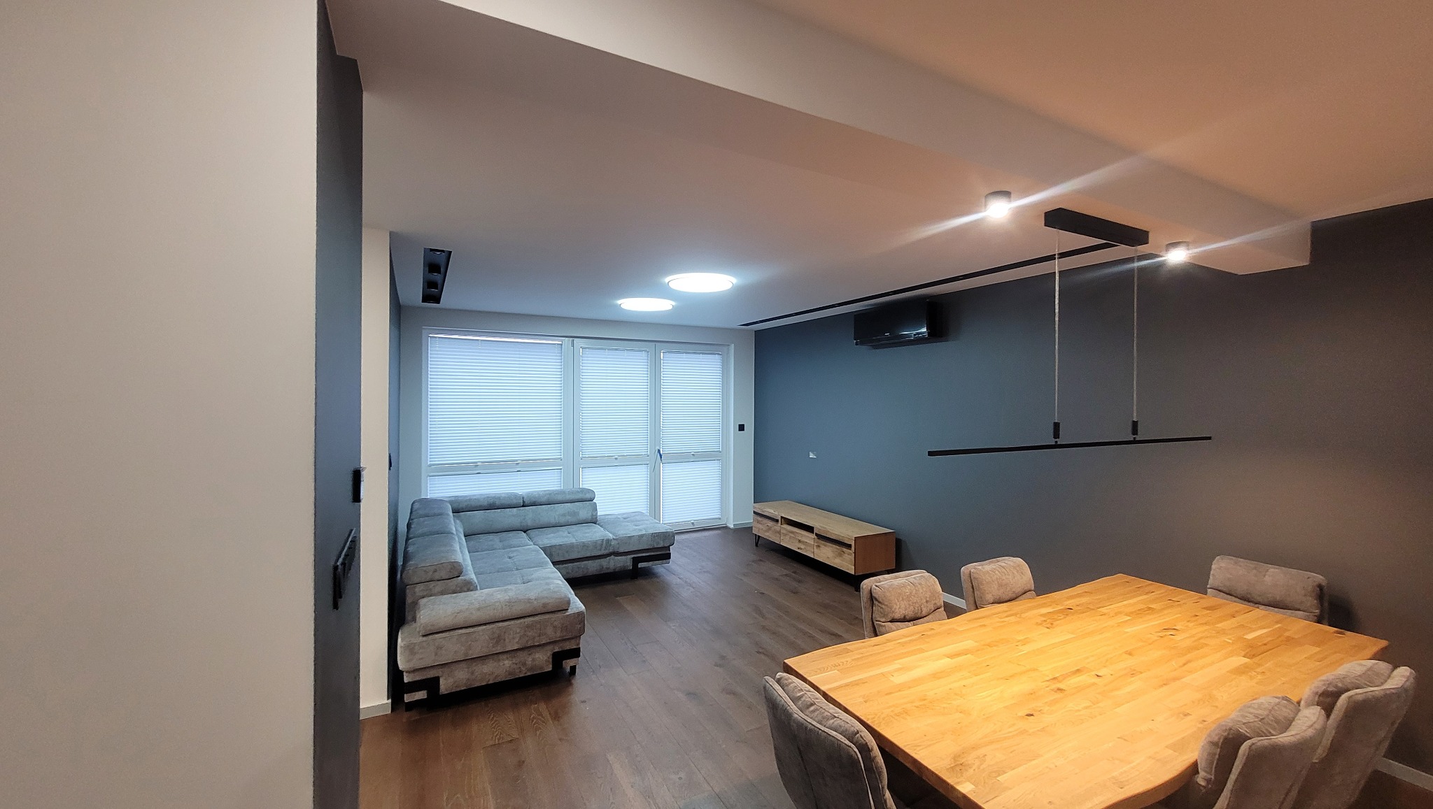 Nowoczesny salon z drewnianą podłogą i stołem, szarą sofą narożną, minimalistycznym oświetleniem i roletami plisowanymi w oknach.