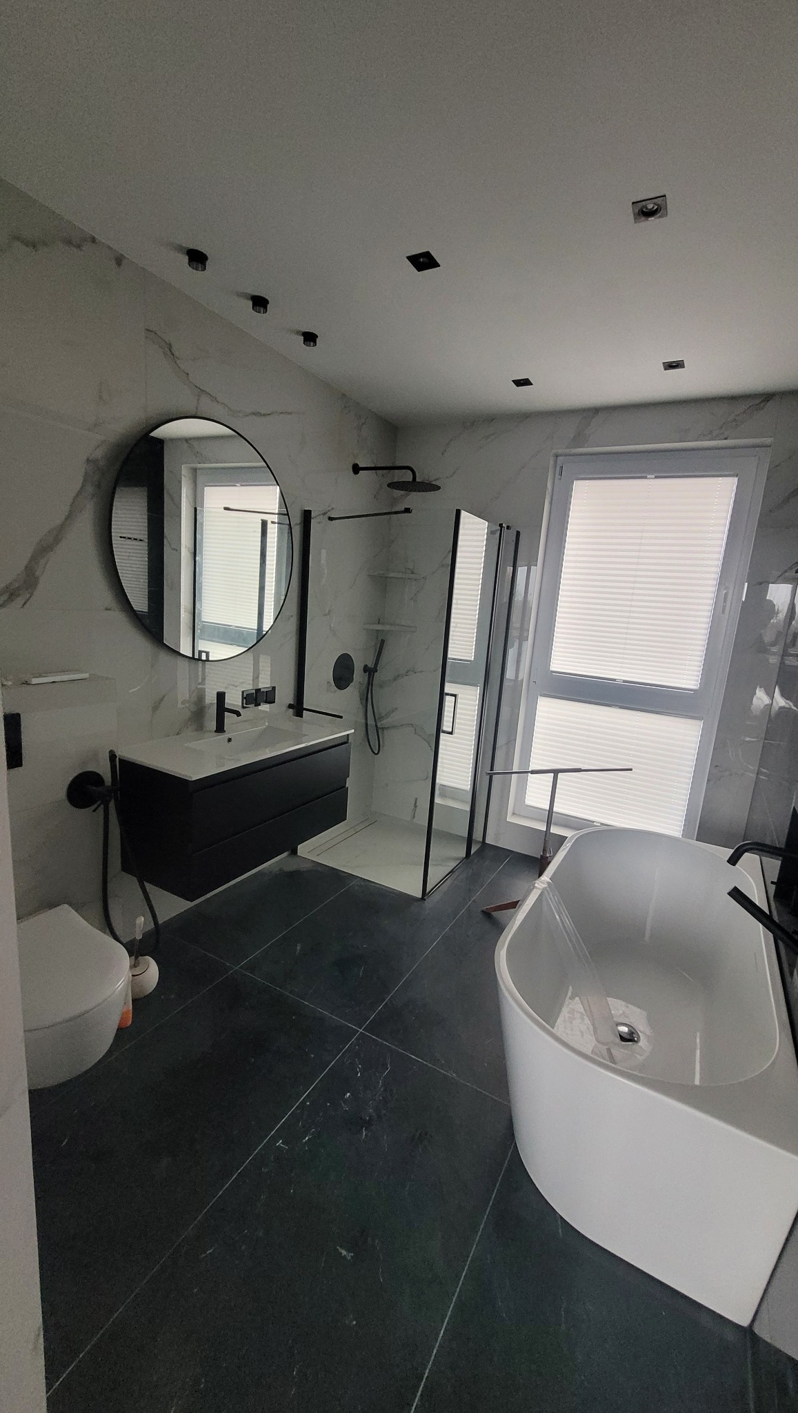 Nowoczesna łazienka w czerni i bieli z okrągłym lustrem, kabiną prysznicową typu walk-in i wolnostojącą wanną, oświetlona punktowymi lampami sufitowymi.