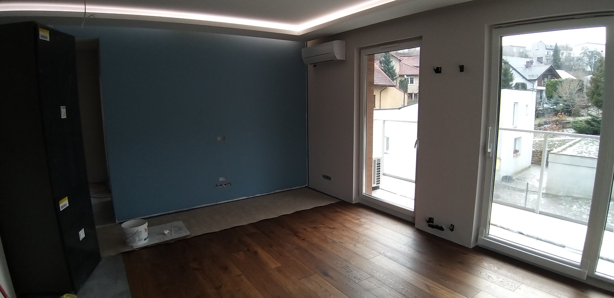 Wnętrze pokoju z drewnianą podłogą i pomalowaną na niebiesko ścianą, widok na zewnątrz przez okno balkonowe, biała listwa LED przy suficie, klimatyzator na ścianie. Na podłodze stoi wiadro po farbie.