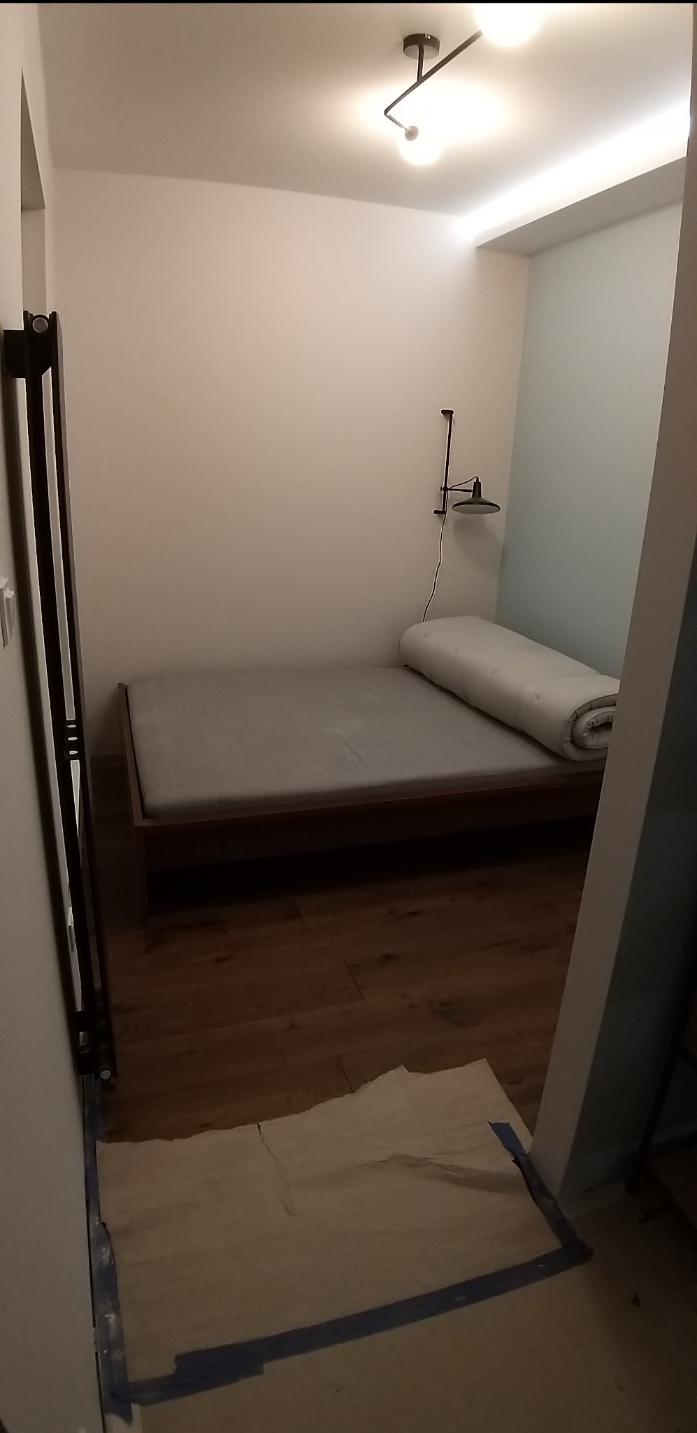 Pusta sypialnia z drewnianą podłogą i materacem na łóżku, zabezpieczona folią malarską przy ścianie, oświetlona lampą sufitową i ścienną.