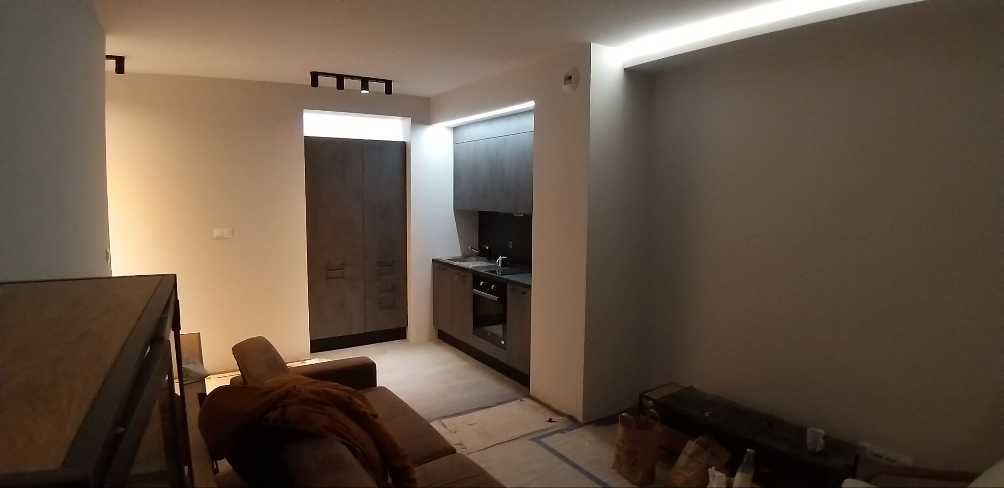 Częściowo wykończone wnętrze mieszkania z szarą zabudową kuchenną, widoczny fragment sofy i zabezpieczona podłoga przed położeniem paneli.