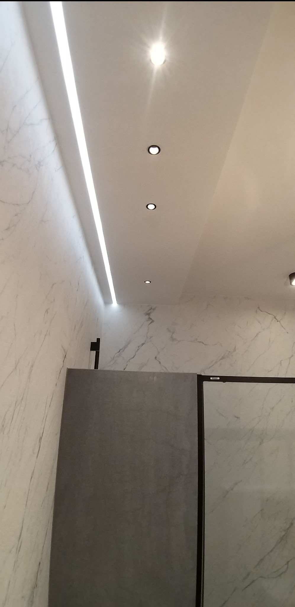 Nowoczesna łazienka z marmurowymi ścianami, oświetleniem LED i punktowymi lampami sufitowymi, widoczny fragment szklanej kabiny prysznicowej.