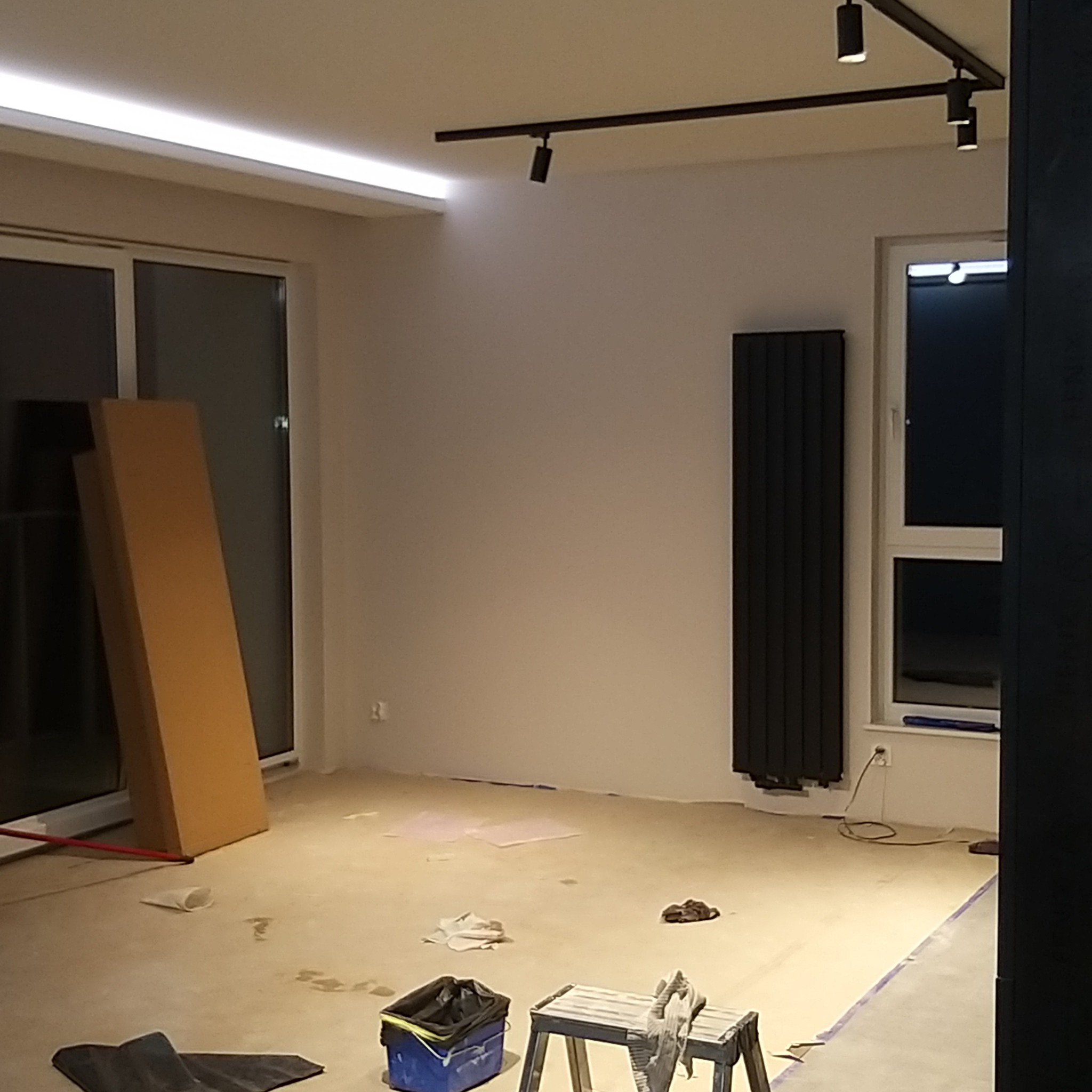 Wnętrze w trakcie remontu: widoczne zabezpieczone okna, grzejnik na ścianie, kartony oparte o ścianę, lampy sufitowe i narzędzia na podłodze.