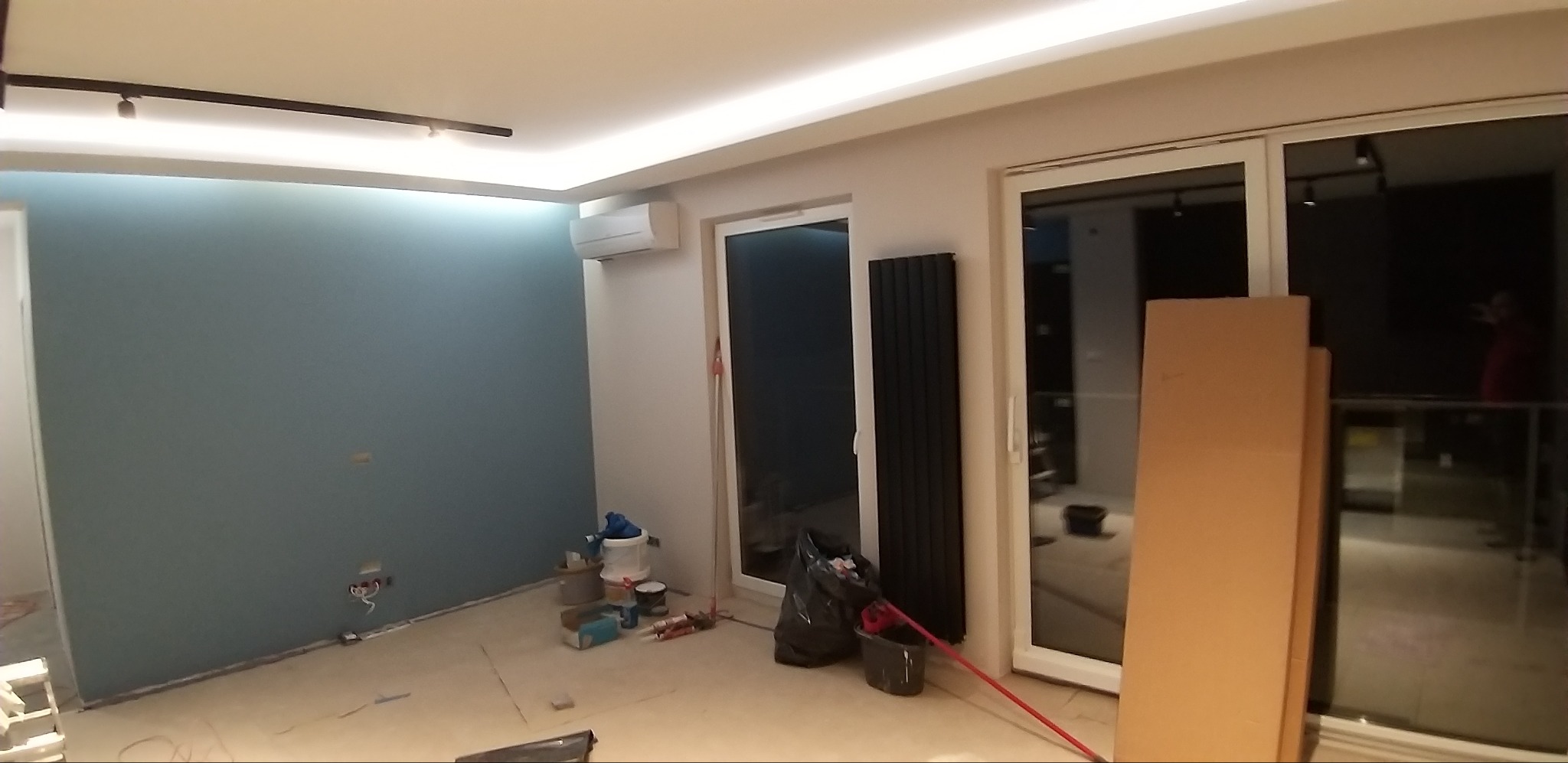 Wnętrze w trakcie prac wykończeniowych: pomalowana na niebiesko ściana, widoczne puszki z farbą, grzejnik panelowy oraz kartony oparty o przeszklone drzwi balkonowe.