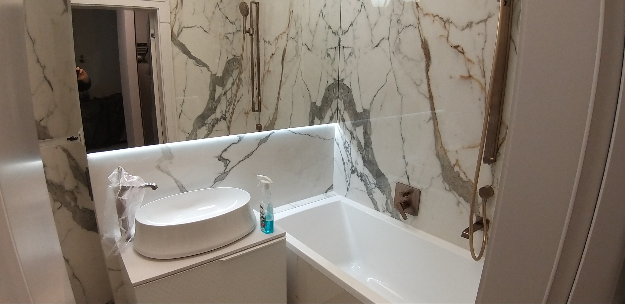 Nowoczesna łazienka z marmurowymi płytkami, owalną umywalką na blacie i wanną, oświetlona listwą LED pod lustrem.