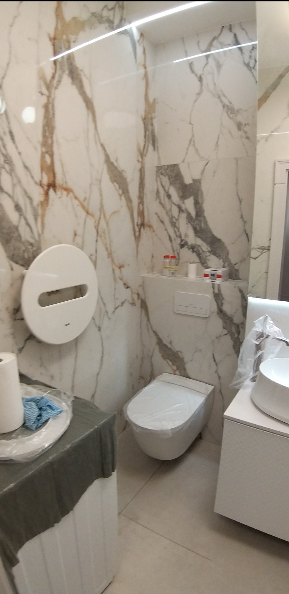 Nowoczesna łazienka z marmurowymi płytkami, wiszącą toaletą i umywalką na szafce, zabezpieczona folią ochronną przed zabrudzeniami podczas prac wykończeniowych.