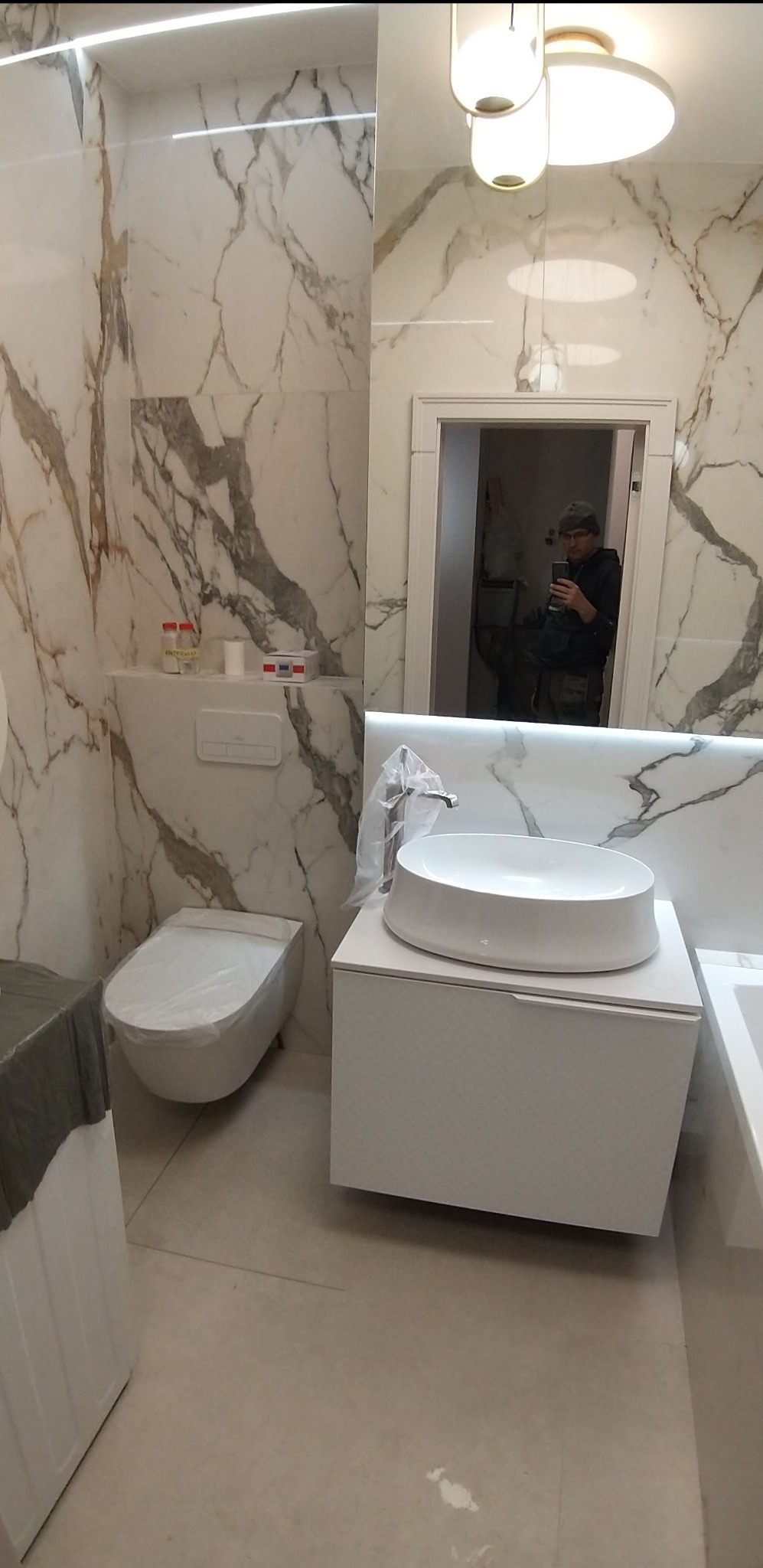 Nowoczesna łazienka z marmurowymi płytkami, owalną umywalką na szafce i toaletą w folii ochronnej, odbicie osoby robiącej zdjęcie w lustrze.