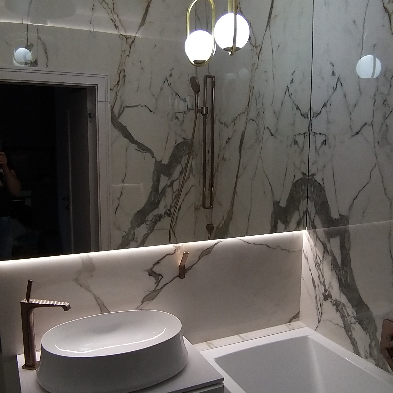 Nowoczesna łazienka z umywalką nablatową, baterią w kolorze różowego złota i marmurowymi płytkami na ścianach, oświetlona lampami kulami.