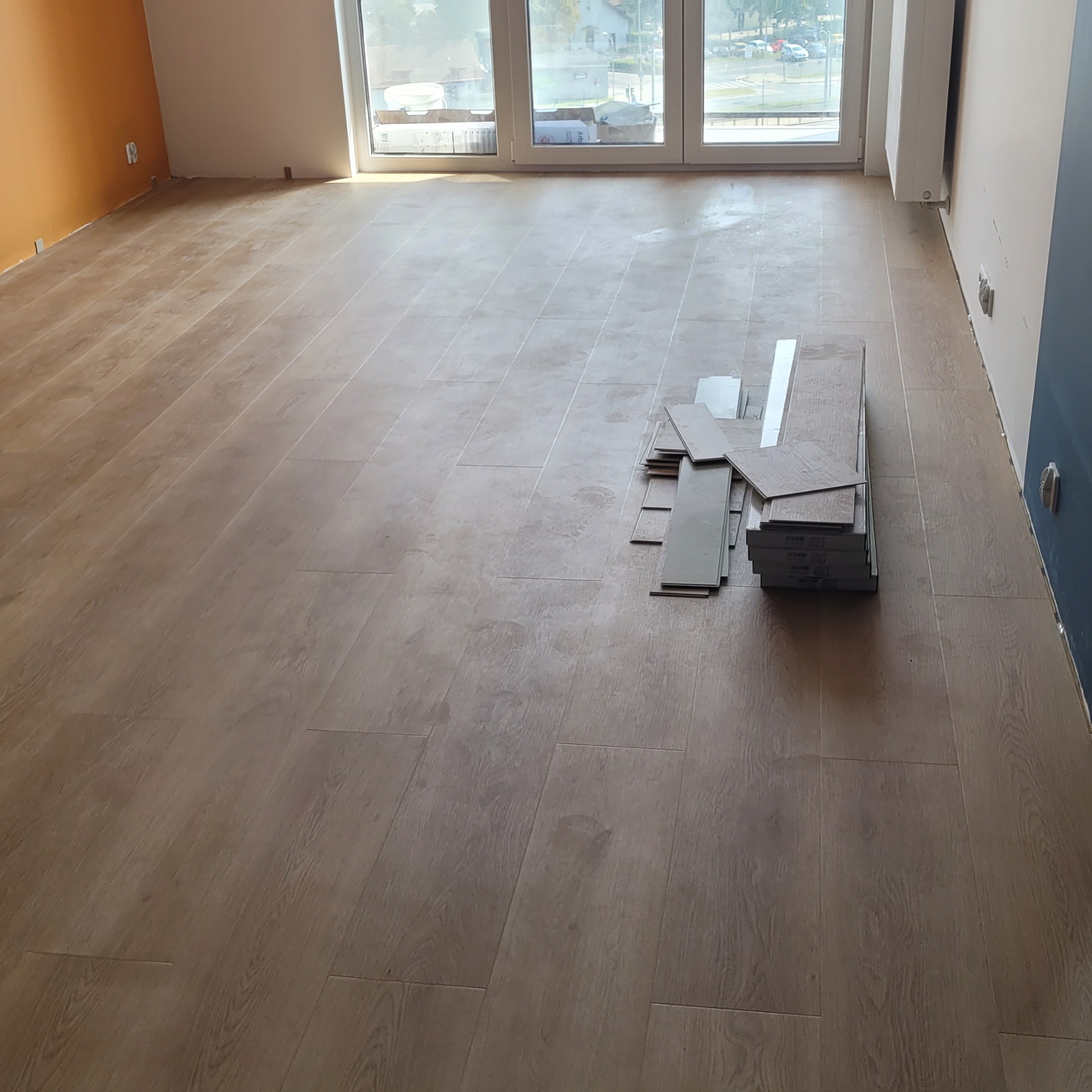 Układanie jasnobrązowych paneli podłogowych w pokoju z pomarańczową i niebieską ścianą, widoczne paczki paneli obok częściowo ułożonej podłogi.