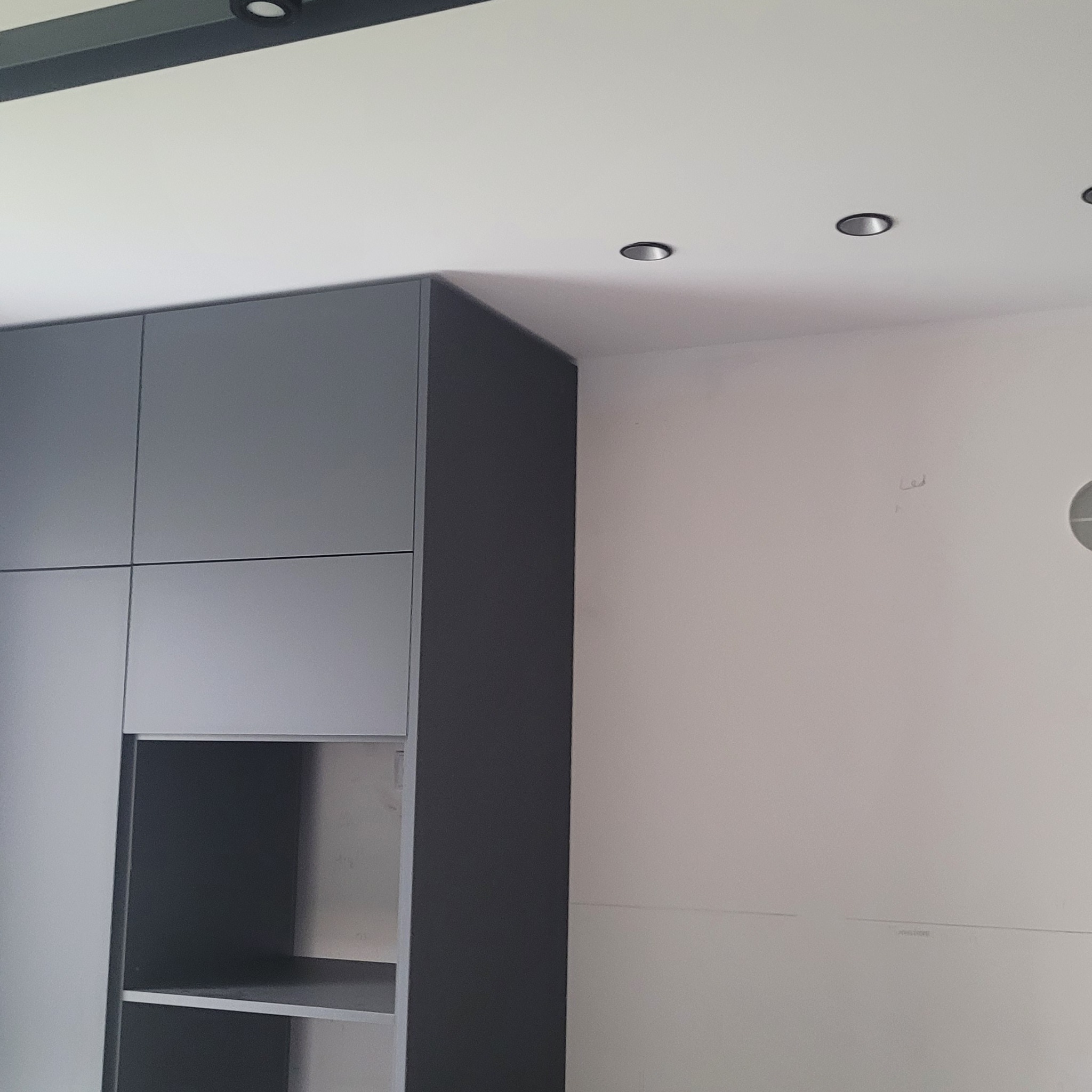 Szara zabudowa meblowa z otwartą półką, w tle biała ściana z widocznymi śladami po pracach wykończeniowych i punktowe oświetlenie sufitowe.