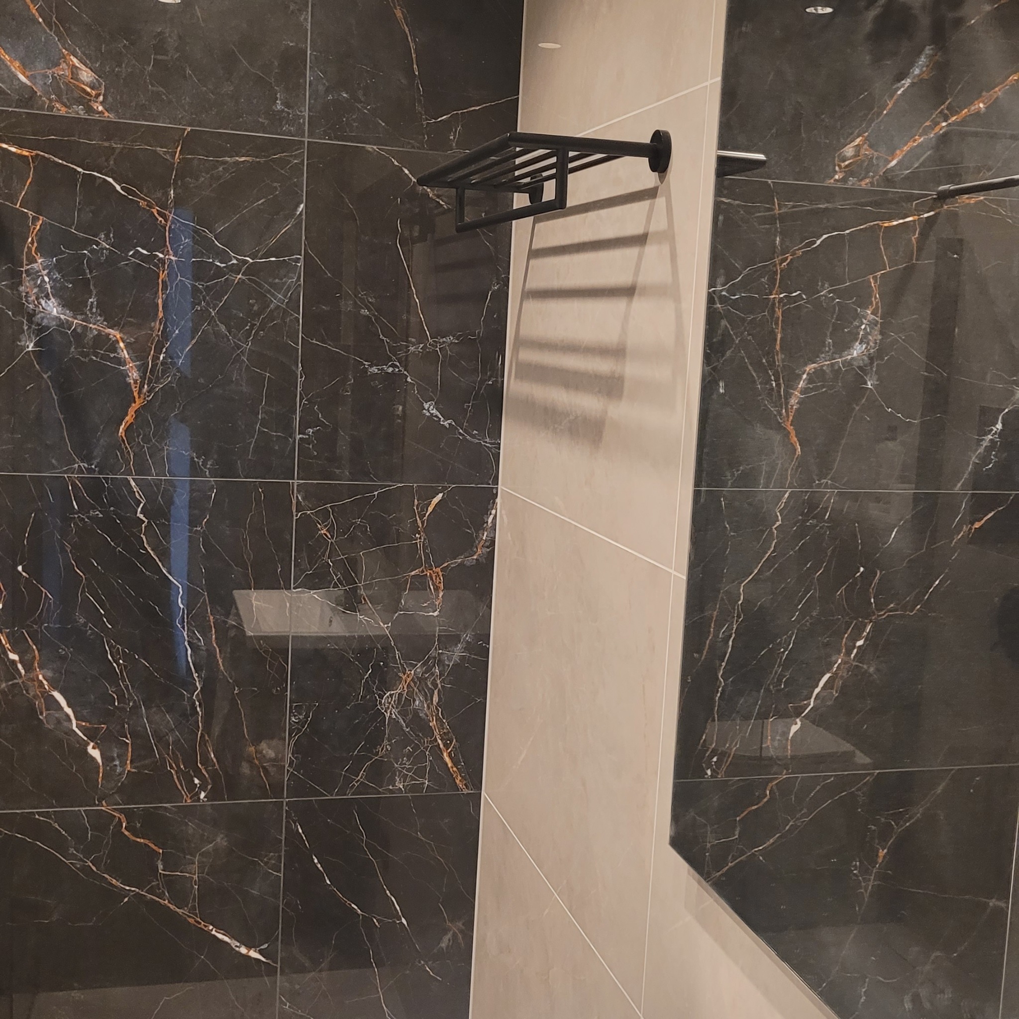 Ciemne płytki imitujące marmur z wyraźnym użyleniem w odcieniach bieli i brązu, zestawione z beżowymi płytkami oraz czarnym, metalowym wieszakiem na ręczniki.
