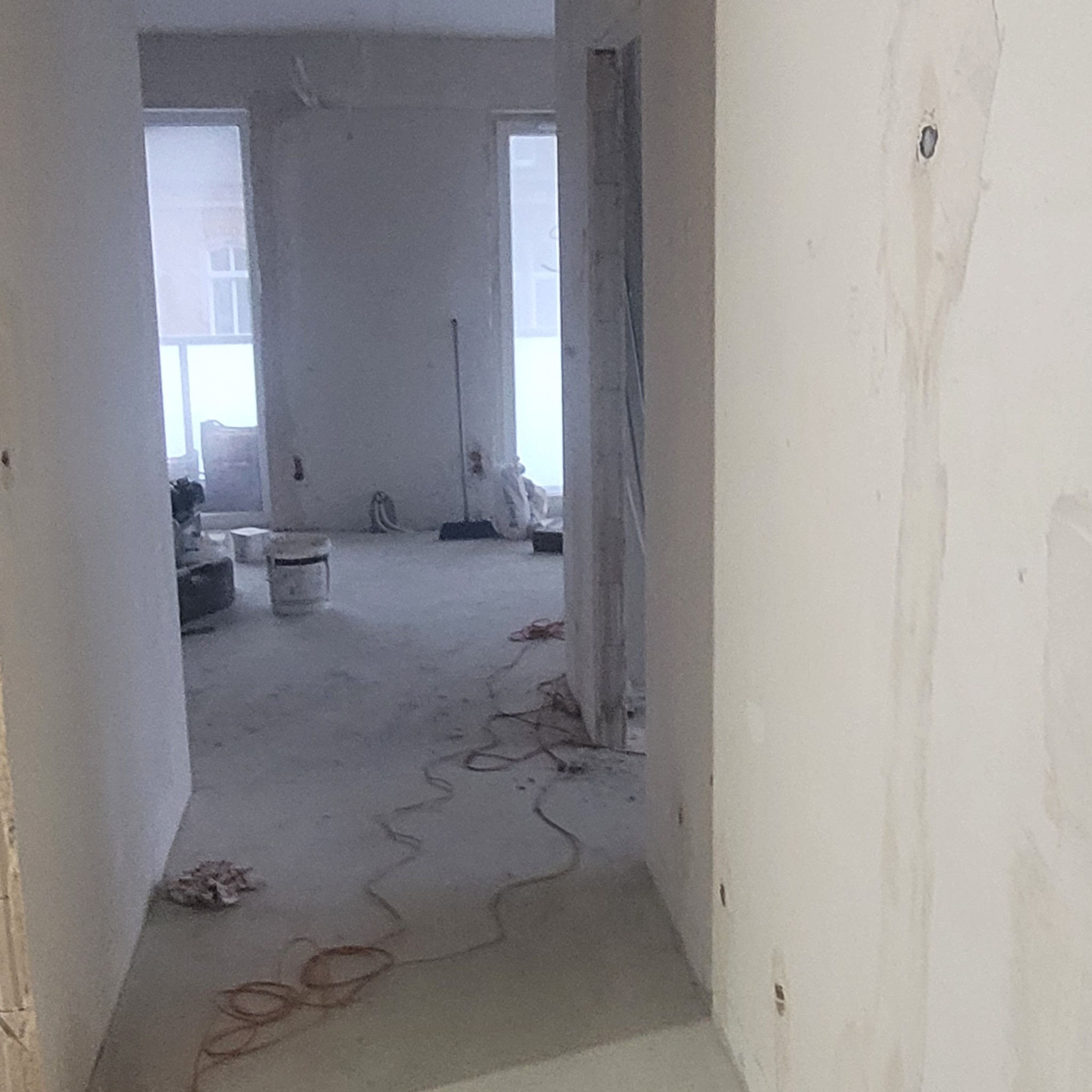 Wnętrze w trakcie prac wykończeniowych: widok na surową ścianę z widocznymi śladami po szpachlowaniu, przewody elektryczne na podłodze oraz wiadro z zaprawą w głębi pomieszczenia.