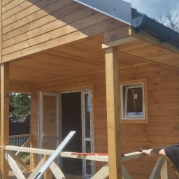 Budmat - Drewniany domek z werandą w trakcie budowy, widoczne otwarte okna i drzwi, poziomica na balustradzie, mężczyzna opierający się o balustradę, błękitne niebo w tle.