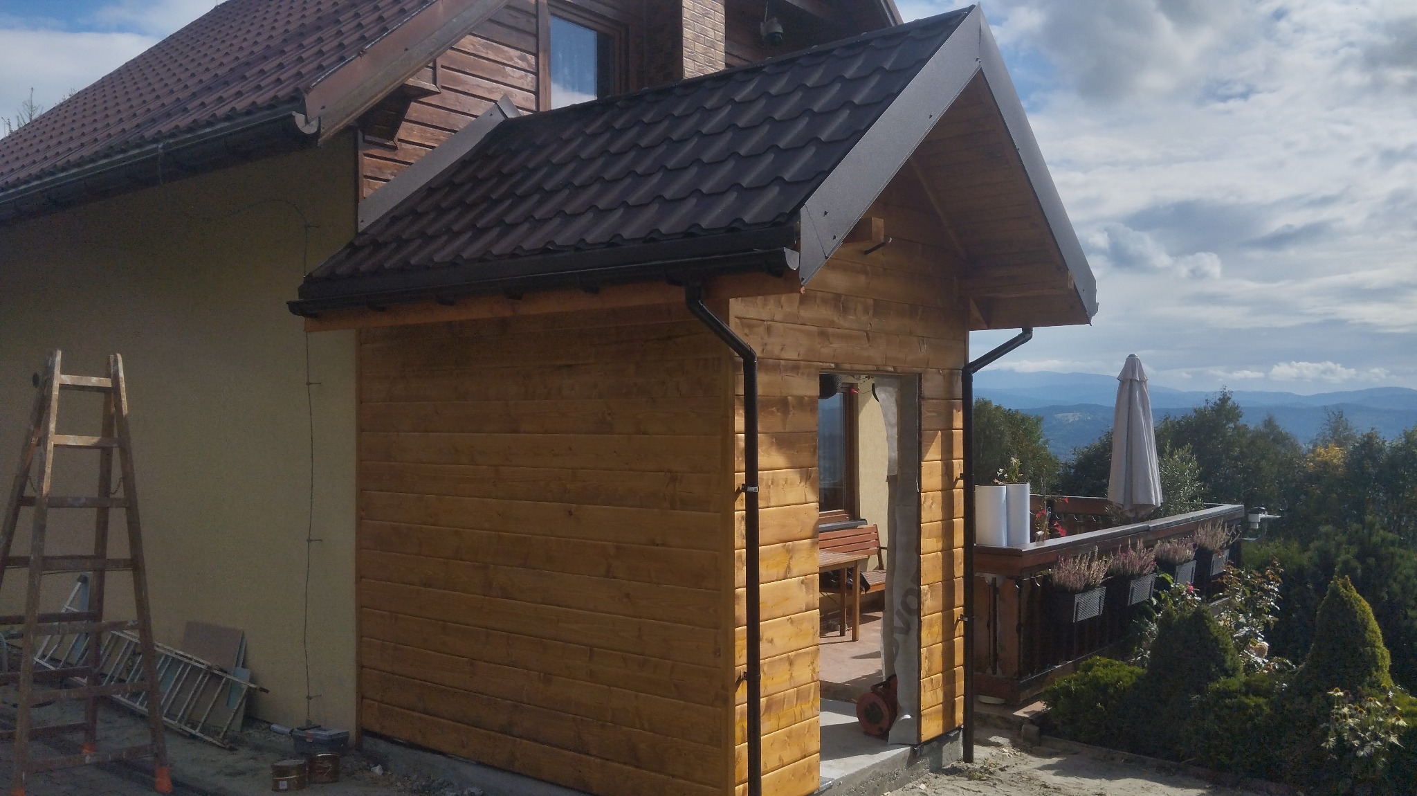 Drewniana dobudówka z ciemnym dachem i rynnami, widoczna drabina, w tle góry i balkon z parasolem.